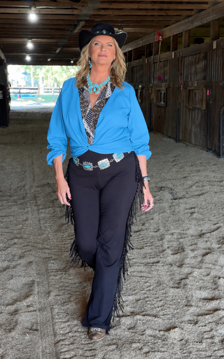 A woman walks down a sandy indoor arena wearing a black cowboy hat, a bright blue button-up shirt, a patterned top, dark navy fringe pants, and turquoise jewelry.