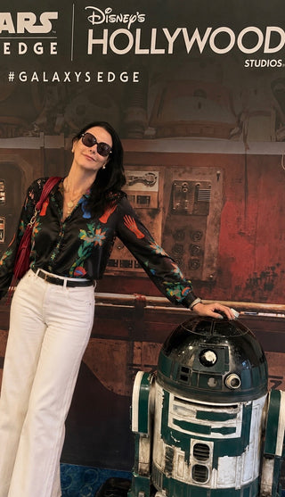 A woman wearing a black floral long-sleeve shirt and white pants stands next to a R2-D2 droid in front of a Disney's Hollywood Studios themed backdrop with Star Wars Edge branding.
