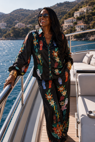 A woman wearing a black satin set with a colorful charms Taboo 2 piece set with mystical charms and hands stands on a yacht deck with blue water and hills in the background.