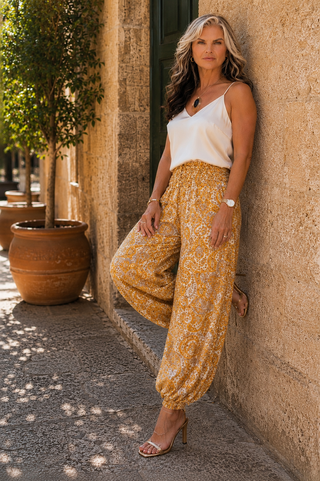 A woman stands on a sunlit stone walkway beside a textured beige wall, wearing a white camisole top and loose, high-waisted yellow-and-white paisley harem pants with clear heeled sandals.