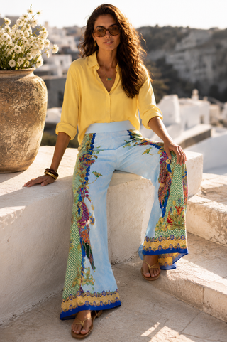 A woman wearing a yellow button-up shirt and light blue wide-leg colorful peaock design pant sitting on white stone steps outdoors, with a sunlit Mediterranean-style background.