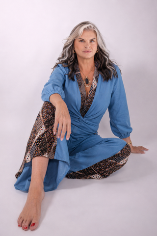 A woman with gray hair sits cross-legged on a light gray studio background, wearing a blue wrap-style robe or pajama set with brown and black patterned trim.
