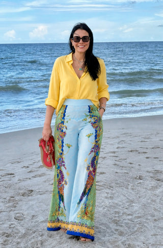 A woman stands on a sandy beach wearing a bright yellow button-up shirt, light blue wide-leg pants with a colorful floral and bird print, and holding a red clutch. The ocean and blue sky are visible in the background.