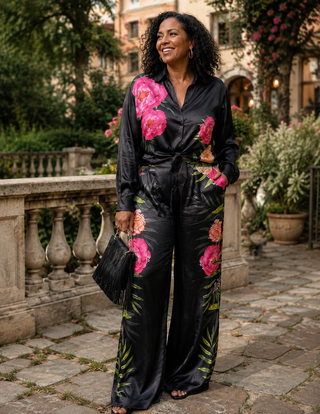 A woman stands outdoors on a stone walkway wearing a black satin jumpsuit with large pink floral prints and green leaves. She is smiling and holding a black handbag.