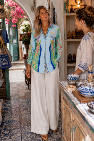 A woman wearing a light blue and green floral embroidered button-up shirt with white wide-leg pants stands in a rustic shop, conversing with another woman.