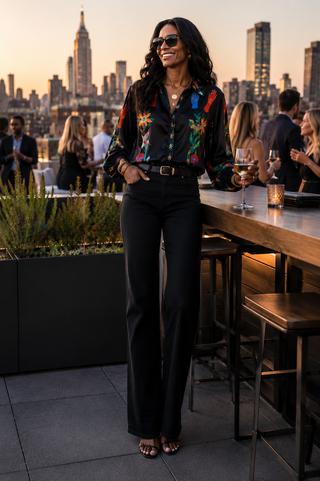 A woman stands on a rooftop bar at sunset wearing a black long-sleeve button-up shirt with a colorful mystical charms and black jeans, with a city skyline in the background.