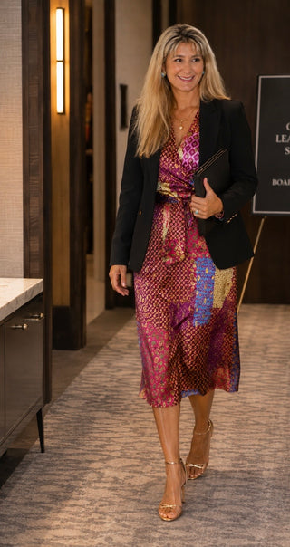 A woman walking in a hallway wearing a black blazer over a vibrant magenta and purple patterned midi dress with gold accents, paired with nude high heels.