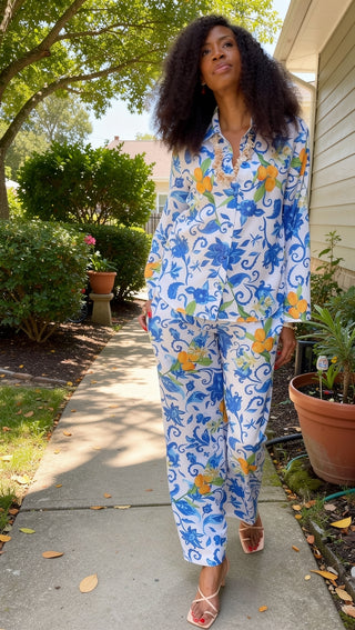 A woman walking outdoors on a garden path wearing a matching two-piece pajama set with a blue and yellow floral pattern on a white background.