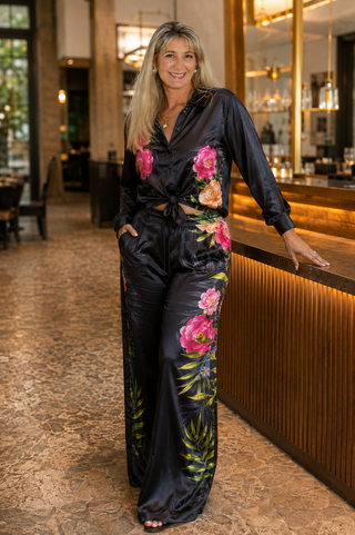 A woman stands in a restaurant or lounge setting wearing a black satin jumpsuit with large pink and peach floral prints and green leaf motifs. The background shows warm wood paneling, a bar counter, and softly blurred interior lights.