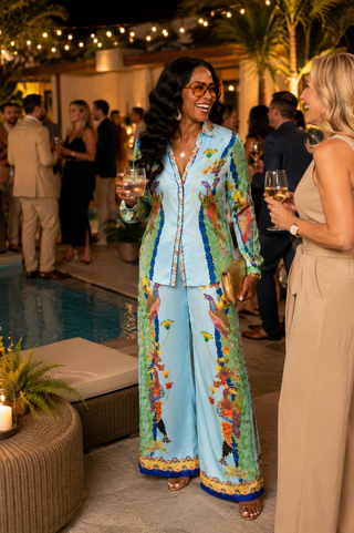 A woman stands by a pool at night wearing a light blue two-piece outfit with colorful tropical bird prints and gold trim, holding a glass of wine in a social outdoor setting.