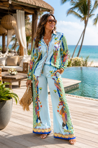 A woman stands on a wooden deck beside a pool wearing a light blue two-piece pajama set with colorful tropical bird prints and gold trim. The set includes a long-sleeve button-front top and matching wide-leg pants.