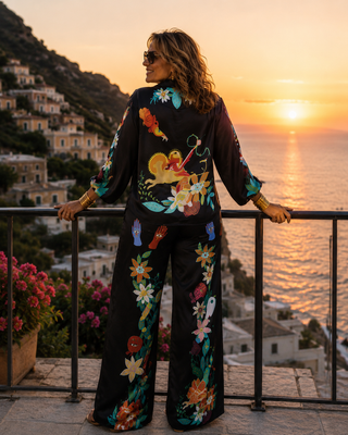 A woman stands on a balcony overlooking a coastal town and sea at sunset, wearing the black mystical charm Taboo satin set.