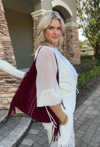 Woman in a white outfit with a burgundy fringed bag standing outdoors.