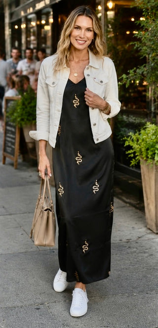 A woman walking outdoors in front of a cafe, wearing a black maxi dress with gold snake prints, a light beige denim jacket, and white sneakers.